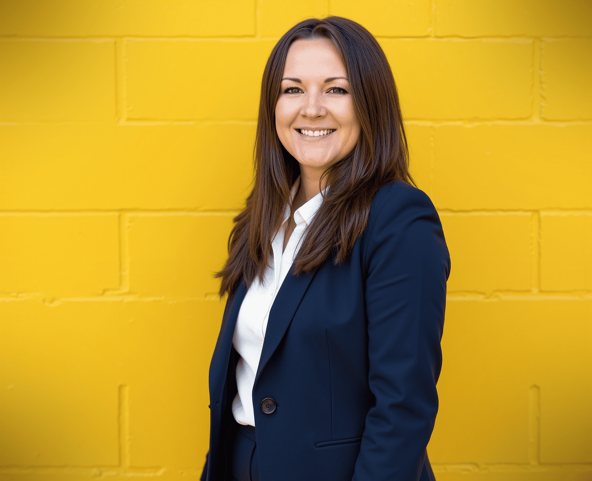 A woman in a navy suit and white shirt stands smiling in front of a bright yellow brick wall.